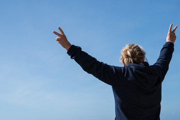 Senior woman with back facing camera holds her arms out making the peace sign against a blue sky. Concept for freedom, retirement, healthy living