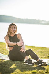 Fototapeta na wymiar Portrait of young sports woman relaxing outdoors after workout sitting on grass on lake background
