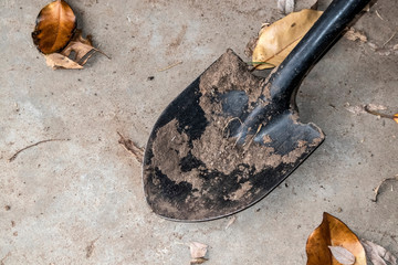 A black shovel with dirt caked on it from digging lays on a sandy concrete surface with brown winter leaves scattered around