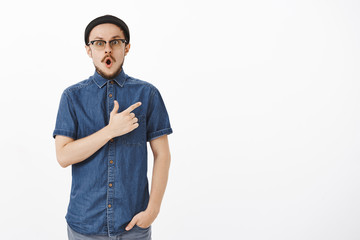 Fototapeta premium Portrait of amazed and impressed handsome young bearded guy in stylish beanie and blue shirt dropping jaw in wow sound pointing at upper right corner staring questioned and thrilled over white wall