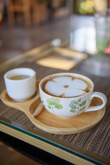 Latte coffee in a white cup, leaf pattern, paired with green tea in a small white cup placed on a wooden plate in a coffee shop.
