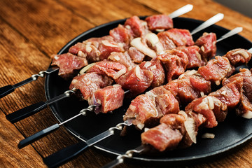 A number of skewers with meat laid out in a black plate. Close-up of mouth-watering pieces of beef ready to fry. Meat dish. Top view on wooden background.