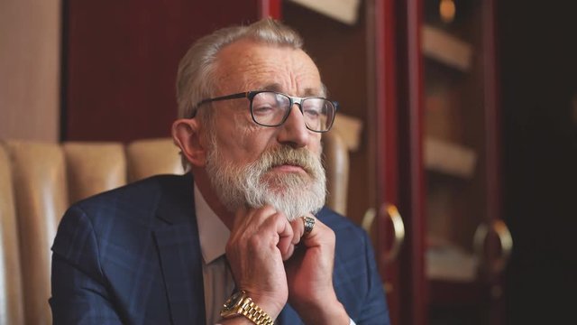 Serious concentrated old male creative artist wears glasses and formal blue suit, with wrinkled and experienced face, remembering remarkable life s moments while sitting in the restautant