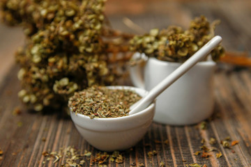 Dried basil spice in a small ceramic bowl, small white ceramic spoon and twig of dried basil on a rustic wooden table.