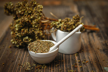 Dried basil spice in a small ceramic bowl, small white ceramic spoon and twig of dried basil on a rustic wooden table.