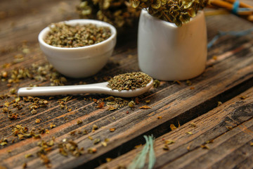 Dried basil spice in a small ceramic bowl, small white ceramic spoon and twig of dried basil on a rustic wooden table.