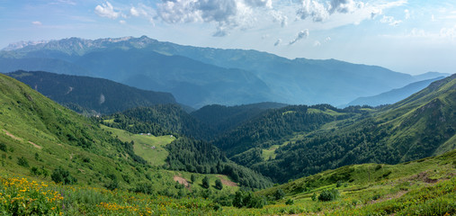 Fototapeta premium The view from the height of a green mountain valley with residential buildings surrounded by high mountains. Snow-capped mountain peaks on the horizon. Krasnaya Polyana, Sochi, Caucasus, Russia.