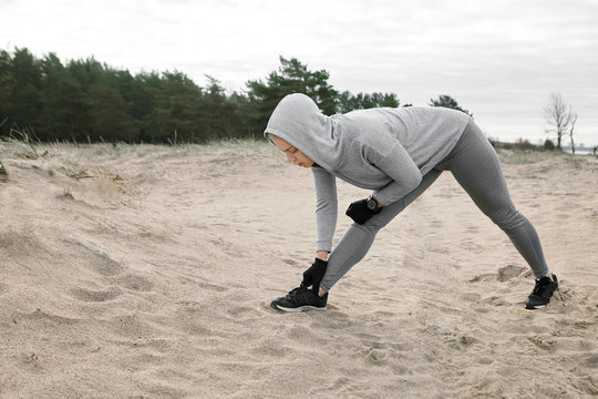 Picture of sporty girl in hoodie, leggings and running shoes warming up leg muscles before cardio workout, standing on sand in chest to knee position. Fit athletic young woman stretching on beach