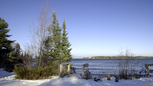 Lake Superior winter with snow and steps leading to shoreline. Conifers, gentle fresh-water waves and lighthouse in background, Upper Peninsula, Michigan. 4K UHD