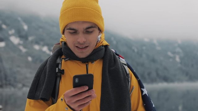 Happy traveler man using smartphone in hiking winter tour. Snowy blurred mountains cape and lake. Traveling and communication concept: hiker scrolling and tapping on line by cell phone