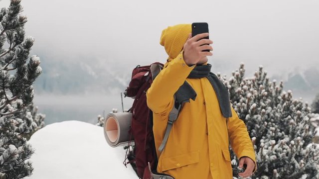 Handsome hiker man video calling on smartphone, waving at camera, smiling and speaking with friends. Beautiful winter mountains and lake against background, Slow motion
