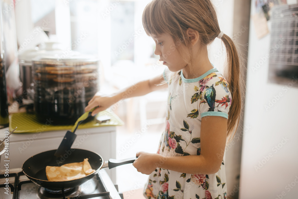 Wall mural little girl is cooking pancakes for the breakfast in the little cozy kitchen