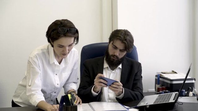 Woman, office worker shows important documents to her bearded boss who is busy with playing smart phone games in the office. Addicted businessman is not listening to his secretary with papers.