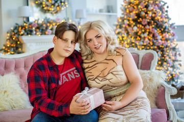 Beautiful mother and her son are sitting together on sofa in cozy room with Christmas tree and fireplace