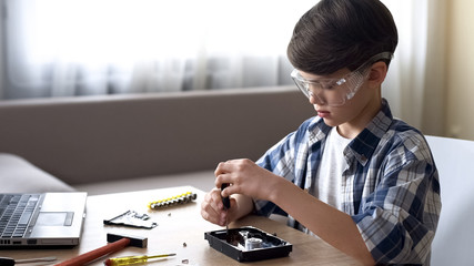 Cute little boy sitting at table and untwisting hard disk drive, IT hobby