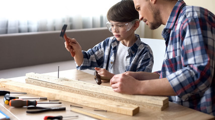 Serious father and son hammering nail in wooden plank, family leisure, support