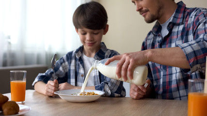 Father pouring milk in plate with corn flakes, son and dad having breakfast