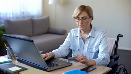 Nice senior woman in wheelchair typing card number on laptop, paying bills