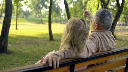 Elderly husband and wife relaxing together in park, man pointing by hand at tree