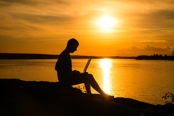 Work on vacation with a laptop computar near the river at beautiful sunset. Handsome man using laptop while sitting by the river and working in the evening.