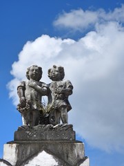 Scene in a cemetery: old stone statue of two children against a sky with large clouds on a sunny day. Singleness and drama.