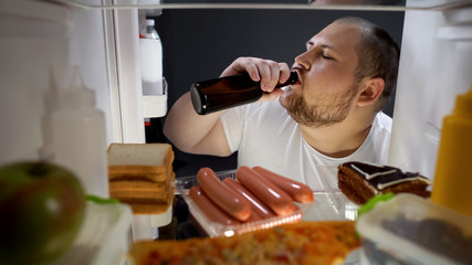 Fat man drinking beer with pleasure from fridge at night, unhealthy lifestyle