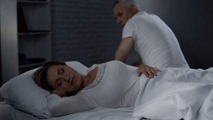 Senior man looking at sleeping wife, male sitting on far bed edge backwards