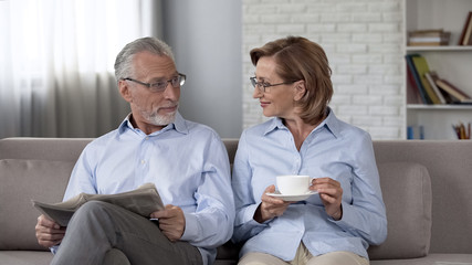 Retiree male and female sitting on sofa man reading newspaper woman enjoying tea