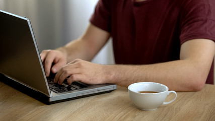 Male freelancer working on laptop and drinking tea at home, student studying
