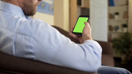 Man with beard sitting on sofa holding green screen smartphone, business app
