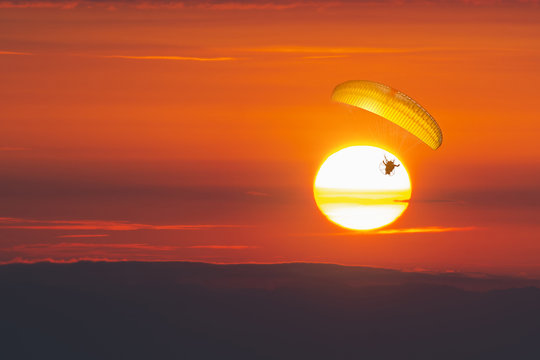 Silhouette of Paramotors flying to sky มParaglider flying with paramotor on dramatic sunset sky with big sun in orange . Concept for adventure extreme ultra light aviation copy space.