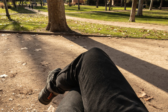 POV Of A Man Legs Crossed Sitting On A Bench In An Outdoor Park