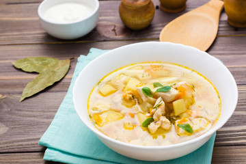 Ready-to-eat chicken soup with potatoes and herbs in a white bowl and sour cream on a wooden table