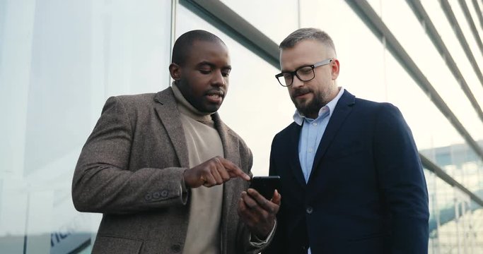 Portrait shot of the two mixed races businessmen talking and one showing another something on the smartphone device screen outdoor the urban office building.