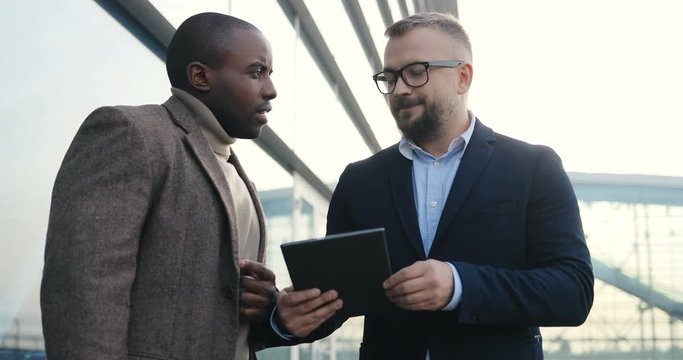 Cauacsian businessman showing something to the African American young handsome man on the tablet device while they standing outside the office center. Portrait.