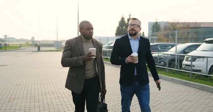Two mixed-races stylish good looking businessmen walking at the parking and talking with coffee in hands. Outdoor.