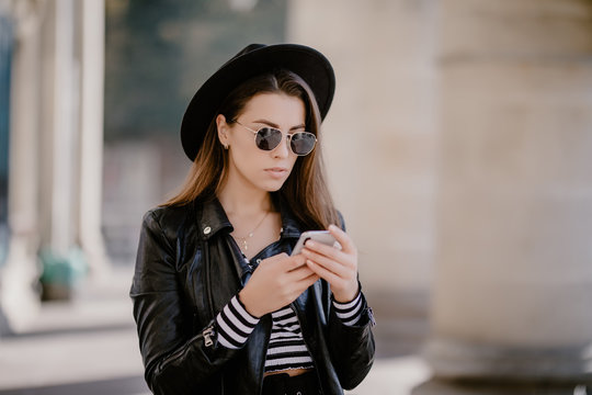 Young pretty Girl in hat and sunglasses checking her smart phone walking in the street outdoors