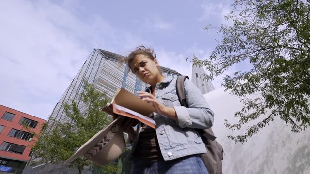 Bottom view of young female reading notes in notebook while standing and go away on background sky and modern buildings in university . Leipzig, Germany