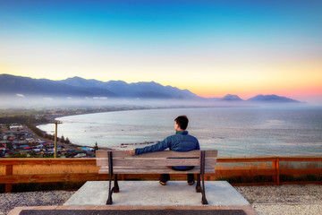 One lonely young man sits on a bench on the top of a hill. The view is breathtaking. There are sunshine, colorful sky, town and coastline and ocean. He is deep thinking and contemplating in his mind.