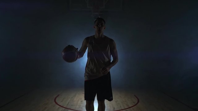 The basketball player goes to the camera and knocks the ball on the ground then stops and holds the ball looking at the camera