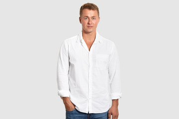 Waist up shot of attractive male entrepreneur wears white shirt and jeans, keeps hand in pocket, isolated over white background. Prosperous young businessman in elegant clothes comes on meeting