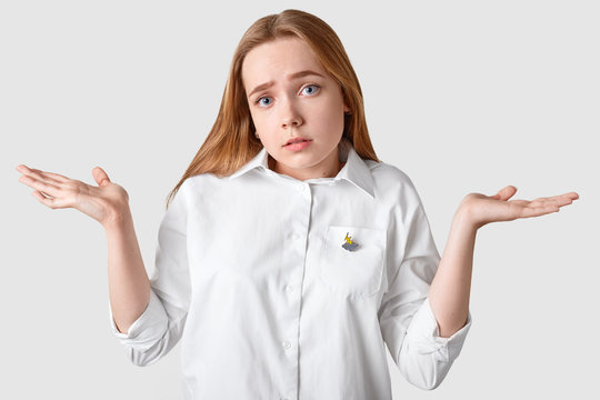Upset clueless girl shruggs shoulders, has indignant facial expression, cant make decision, looks confusingly at camera, wears elegant shirt, isolated over white background. Unaware youngster indoor