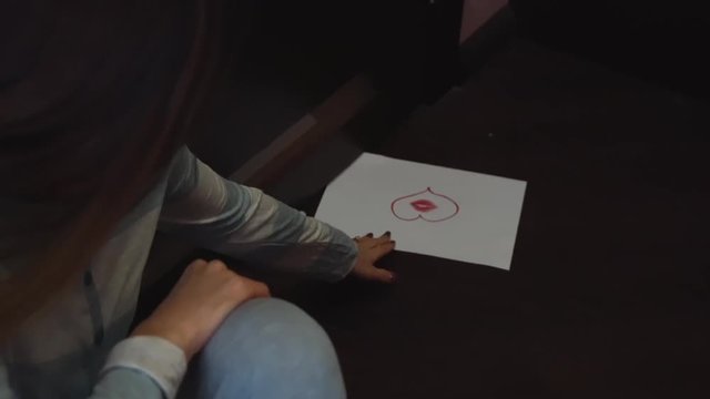 Sad woman sitting on a floor in dark room and taking a sheet of paper with painted heart, which have appeared under the door. Close-up.
