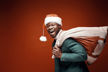 Modern Santa Claus. Smiling emotional man posing in green coat and red sweater, with santa hat and bag. Studio shot, brown background