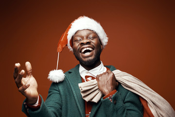 Modern Santa Claus. Smiling emotional man posing in green coat and red sweater, with santa hat and bag. Studio shot, brown background