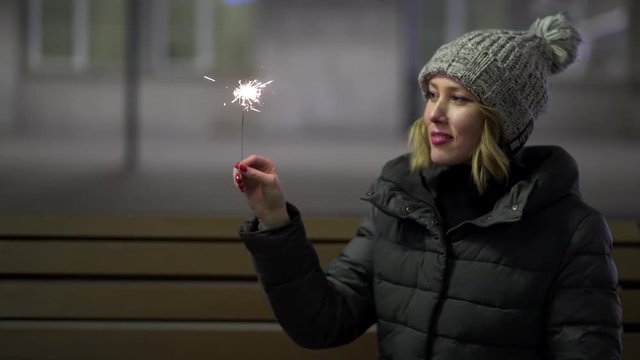 Smiling, beautiful girl in knitted hat and down jacket in the street at night with sparkler, celebrating New year, Merry Christmas. Young, pretty woman holding bengal fire on grey street background.