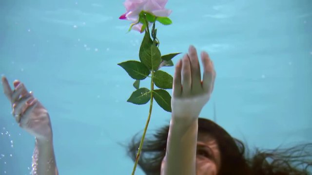 Close-up portrait of a charming brunnete woman catching floating flower under the water, smiling princess in a white dress with a beautiful flower.