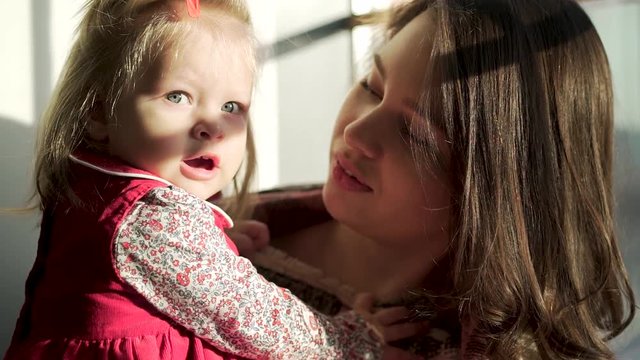Beautiful baby girl with cute chubby cheeks looking straight in her mother arms. Lovely child and her mom against the window enjoy while spending time together.