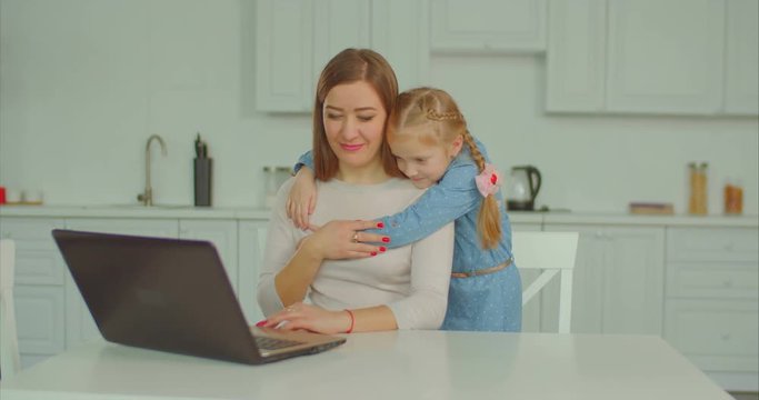 Affectionate caring elementary age daughter embracing her busy overworked mother from behind with love while freelance woman networking on laptop pc in domestic kitchen.