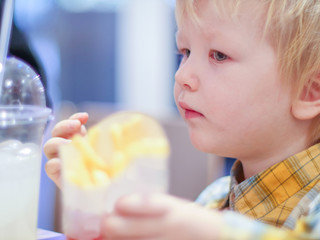 Cute little blond child drinks carbonated drink through straw and eats fried potato in special fast food cafe. Boy likes to eat among people. Concept of fast food. Shallow focus, blur.